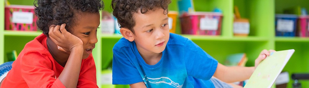 Dos niños leen un libro recostados en el piso del aula.